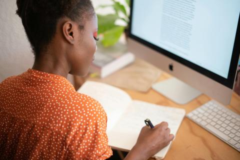 A behind-the-shoulder shot of a woman at her desk writing in a notebook
